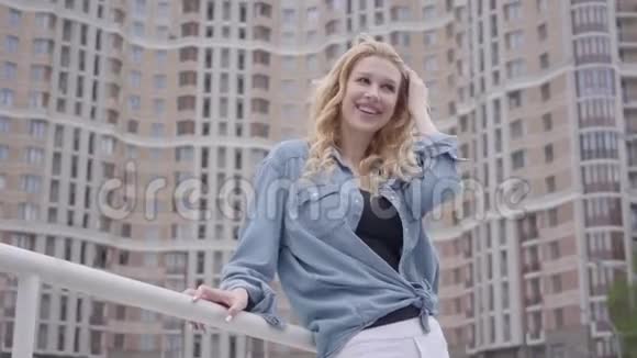 漂亮自信的金发女人穿着牛仔裤夹克摆在摩天大楼前微笑着看着远处城市生活方式城市视频的预览图