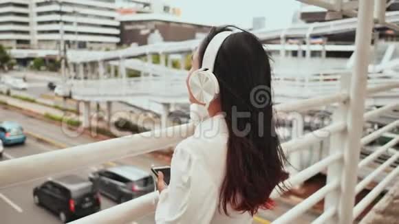 一个亚洲女孩站在交通桥上戴着白色耳机听音乐视频的预览图