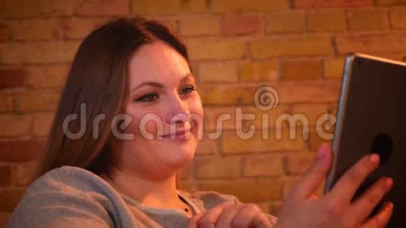 超重快乐女模特的特写肖像坐在沙发上在舒适的家庭氛围中微笑着走进平板电脑视频的预览图