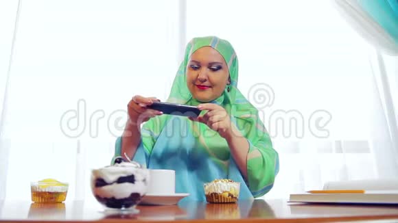 一个年轻的穆斯林妇女戴着绿色围巾在咖啡馆里拍摄电话上的糕点视频的预览图