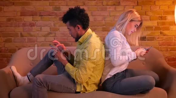 非洲男孩和白人女孩背靠背地坐在沙发上看着家里的智能手机视频的预览图