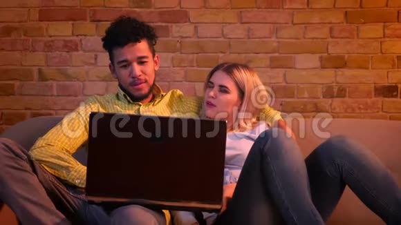 年轻的非洲男人和高加索女孩坐在沙发上手提电脑在家里愉快地交谈视频的预览图
