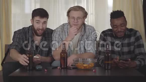 三个快乐的男人朋友坐在家里看着电视喝着带薯片的啤酒有一个视频电话给一个视频的预览图