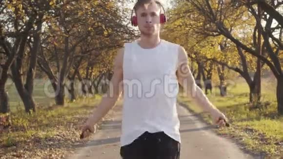 年轻的白人男孩戴着耳机和运动服在秋天公园跳绳户外运动运动员训练视频的预览图