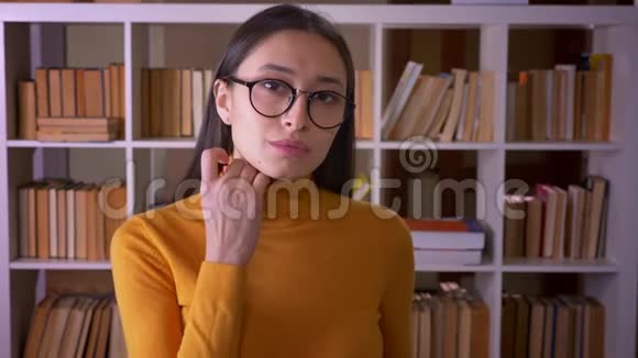 性感的黑发女教师的肖像戴着眼镜摆成相机在图书馆调情视频的预览图