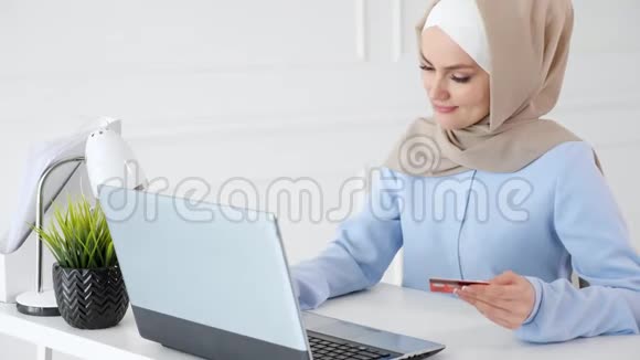 戴着头巾的穆斯林妇女正在用信用卡和笔记本电脑在线购买视频的预览图