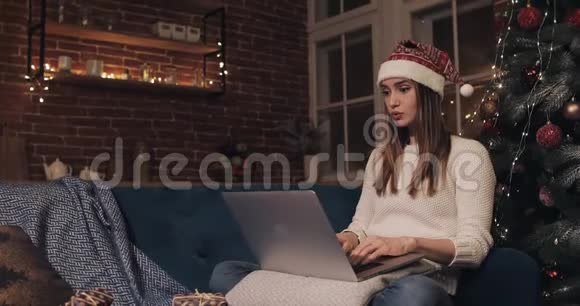 漂亮的年轻女子戴着桑塔帽坐在家中圣诞树附近背景是她用笔记本电脑发短信打字视频的预览图