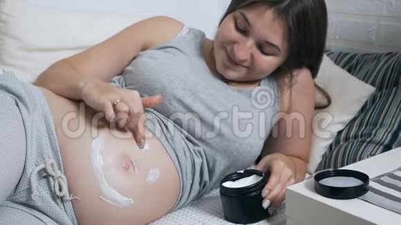 幸福的孕妇躺在家里的沙发上用面霜涂抹在肚子上露出笑容视频的预览图