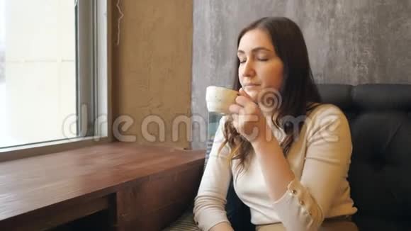 美丽的年轻女子在咖啡馆的窗户附近喝着一杯热咖啡视频的预览图