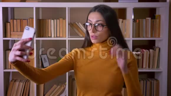 美丽的黑发女教师眼镜制作自拍智能手机在图书馆视频的预览图