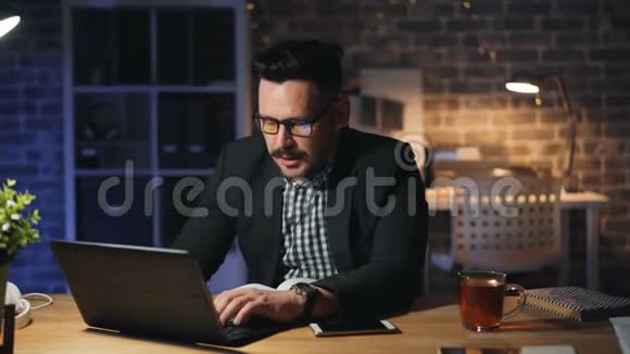 精疲力竭的企业家打哈欠在黑暗的办公室里用笔记本电脑工作的肖像视频的预览图