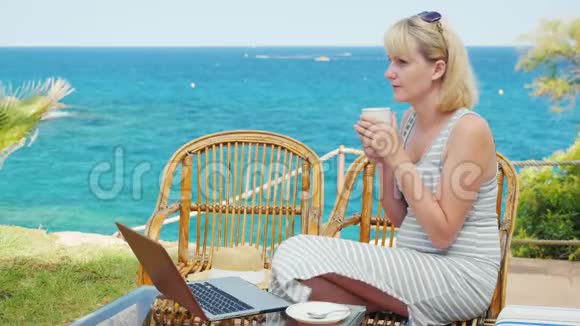喝咖啡的年轻女人坐在露台上俯瞰大海笔记本电脑旁边视频的预览图
