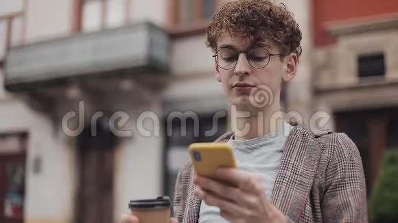 年轻的嬉皮士穿着夹克和眼镜一边喝着咖啡一边在坐着一边嘲笑他的现代手机屏幕视频的预览图