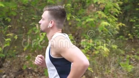初秋一个运动的年轻人沿着森林附近的小径奔跑带无线耳机的帅哥慢跑视频的预览图