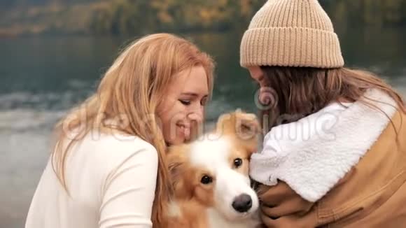 两位年轻时尚的白人女性在山上的湖上与她的边境牧羊犬玩耍和亲吻视频的预览图