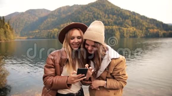 两个迷人的有趣女孩年轻女性穿着时髦的衣服在山上的一个湖上度假看着一个移动电话视频的预览图