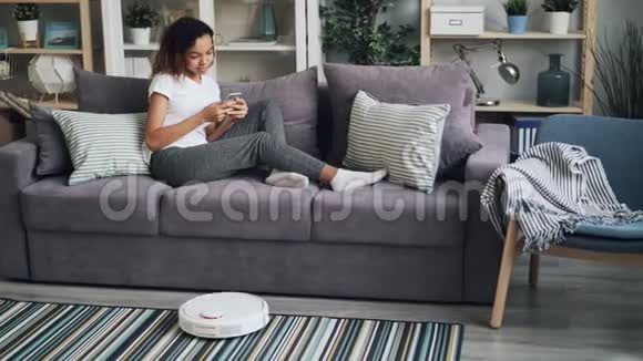 年轻的非洲裔美国妇女正在使用智能手机并在沙发上放松而机器人吸尘器正在吸尘视频的预览图