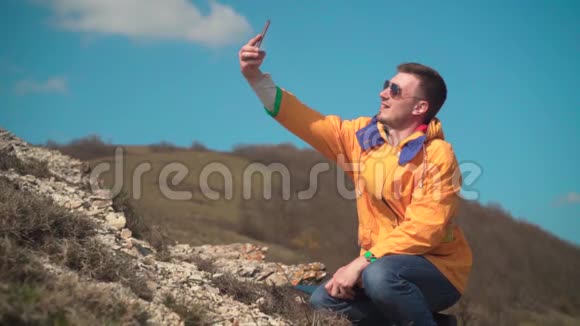 一个穿着黄色夹克蓝色牛仔裤和眼镜的男人坐在山上欣赏风景自拍拍照视频的预览图