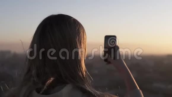 长头发的年轻女子从后面站着在城市模糊的背景下使用手机日落或日出视频的预览图
