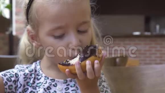 有趣的小女孩在咖啡馆的桌子上吃甜巧克力甜甜圈可爱的金发女孩咬着巧克力甜甜圈视频的预览图