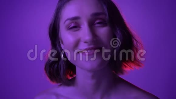 年轻迷人的女性在镜头前用霓虹灯紫色背景诱人微笑的特写镜头视频的预览图