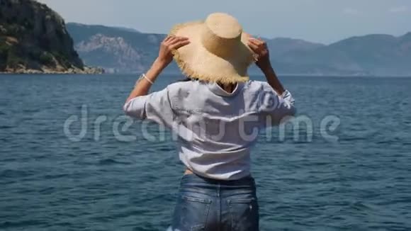 穿着蓝色衬衫和牛仔短裤的女孩戴着草帽站在海边视频的预览图