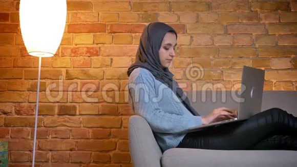 在室内坐在沙发上用笔记本电脑拍摄年轻迷人的穆斯林女性头巾的特写侧景视频的预览图