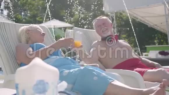 年长的积极夫妇躺在游泳池附近的日光浴床上喝果汁交谈和微笑充满爱意的家庭休息视频的预览图