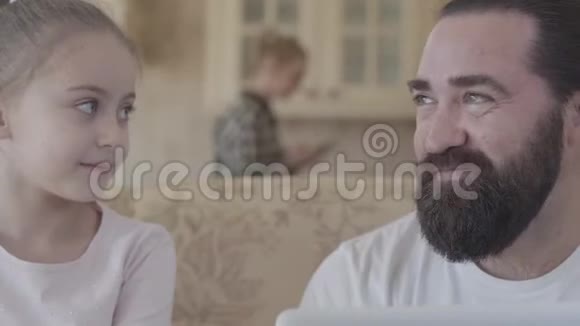 大胡子男人坐在沙发上和他的小女儿近距离交谈背景中母亲模糊的身影视频的预览图