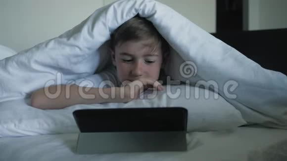 一个男孩的特写镜头是在一张毯子下面的数字平板电脑上看卡通电子成瘾概念视频的预览图