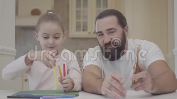 年轻的父亲和他可爱的小女儿坐在客厅里看着用记号笔和投掷的实验视频的预览图
