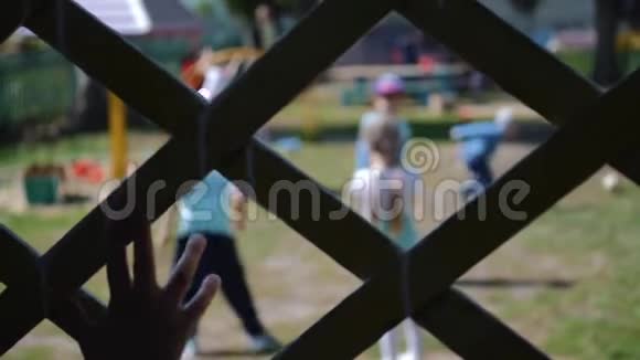 一个孤独的孩子拿着篱笆看着孩子们玩耍宏观的手视频的预览图