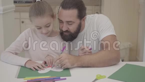 年轻的父亲和他可爱的小女儿坐在客厅里用绿色纸板上的标记明信片画画视频的预览图