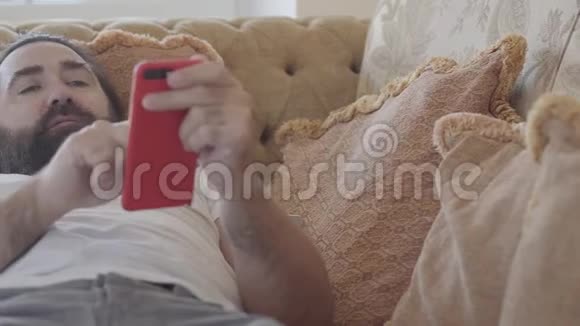 一个留着胡须的人在柔软的米色沙发上玩智能手机游戏在失败和被抛弃之后视频的预览图