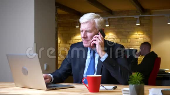 一个老白种人商人在办公室里用笔记本电脑时打电话的特写镜头视频的预览图