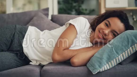 微笑的非裔美国女士躺在沙发上头枕在枕头上看着相机的照片日间休息视频的预览图