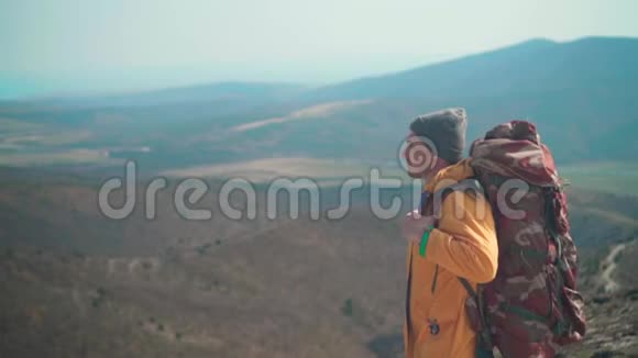 一个穿着黄色夹克蓝色牛仔裤和带着一个大型旅游背包的帽子的年轻人站在山上望着远处视频的预览图