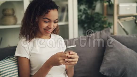 快乐的非裔美国女人拿着智能手机摸着屏幕坐在灯光室的沙发上笑青年视频的预览图