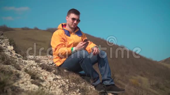 一个穿着黄色夹克蓝色牛仔裤和眼镜的男人坐在山上欣赏风景在电话里说话笑视频的预览图