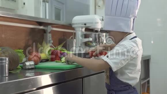 小厨师穿着围裙和厨师帽在学校厨房做饭有趣的小男孩做饭切新鲜蔬菜视频的预览图