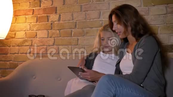 年轻的白种人母亲和她漂亮的金发小女孩坐在平板电脑上的特写镜头视频的预览图