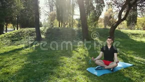 年轻人在公园练习瑜伽户外坐在垫子上坐在莲花姿势上闭着眼睛沉思道具慢动作视频的预览图