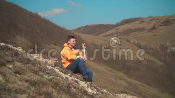 一个穿着黄色夹克蓝色牛仔裤的男人坐在山上透过望远镜看然后在电话里说话视频的预览图