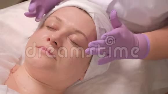 美容师双手戴着紫丁香手套眼睛周围放着奶油盯着中年妇女脸美容治疗程序视频的预览图