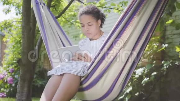 年轻的非洲裔美国妇女坐在吊床上在花园里放松在她的平板电脑上发短信户外休闲视频的预览图