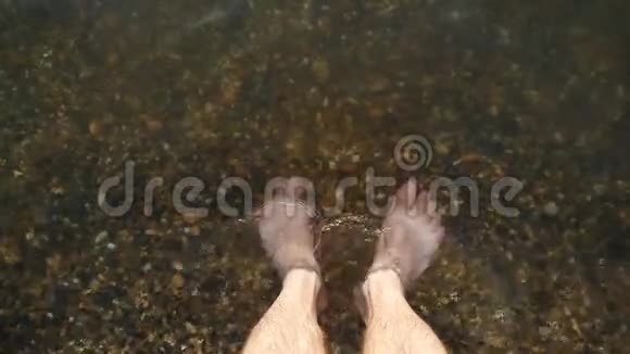 一个光着脚的人站在水中的一个鹅卵石海岸上水在近距离地他的脚视频的预览图