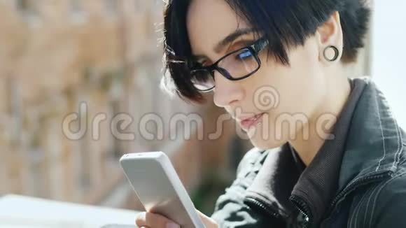 戴着时髦眼镜的年轻女人的肖像它用窗户上的手机做梦视频的预览图