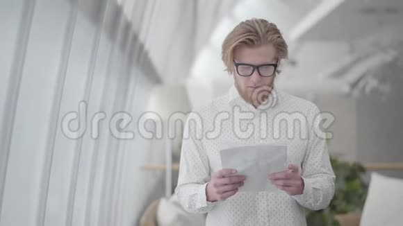 肖像成功的金发男子戴着眼镜站在一个轻松舒适的办公室阅读文件帅哥商人视频的预览图