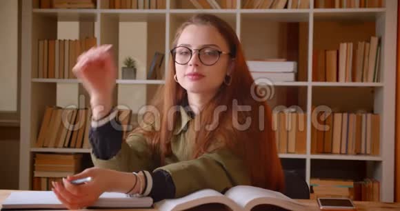 一幅年轻的女学生坐在图书馆里带着书看着镜头视频的预览图