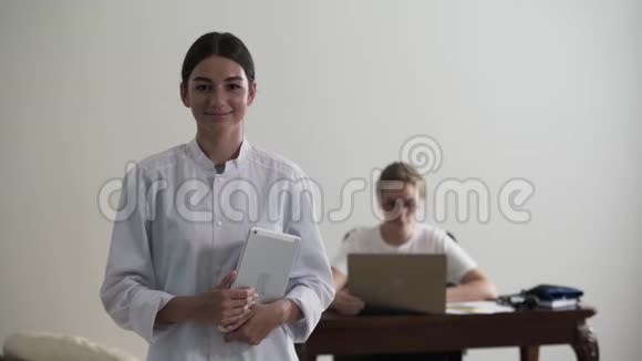 年轻专业女医生拿着笔记本电脑看镜头的肖像在后台坐着一个女孩视频的预览图